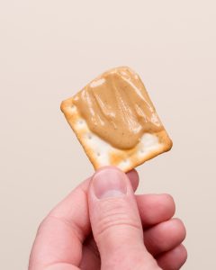 A hand holding a square cracker topped with a generous spread of creamy peanut butter against a plain beige background.