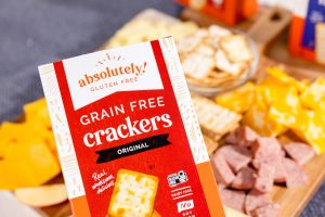 A box of Absolutely! Grain Free Crackers is held in front of a charcuterie board with cheese, meats, and more crackers.